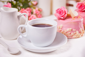 A cup of tea, pink roses and small cake on the white table