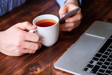 Freelancer relaxes drinking tea and browsing social networks through the phone next to his laptop.