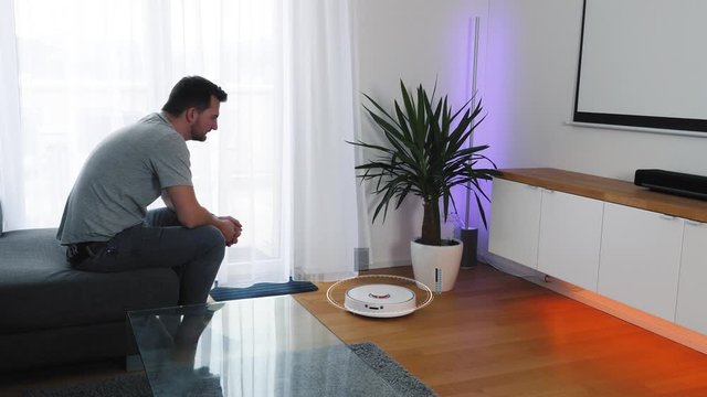 Man sits on a couch in the living room and activates his vacuum robot cleaner via smartphone voice control