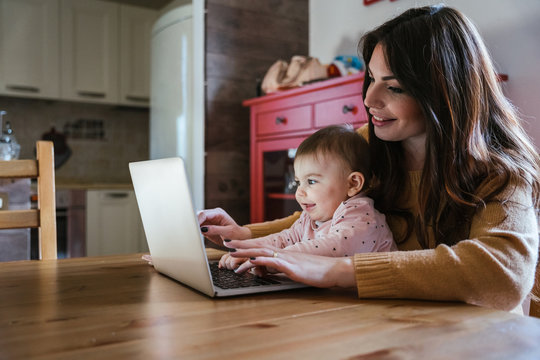 Portrait of a young mom sitting at the kitchen table at home with her child daughter while working on the laptop - Millennial with a little girl and having fun together