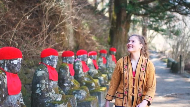 Slow motion backwards shot of young woman walking, looking at row of Jizo statues in Kanmangafuchi Abyss in Tochigi prefecture in Nikko, Japan in early spring with moss