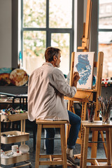 Back view of a senior artist sitting in his studio in front of an easel