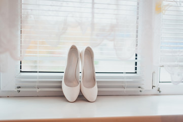 Wedding white leather shoes stand against the background of a window in the interior. Preparation and morning of the bride. Photography, concept.