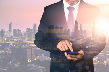 The double exposure image of the business man using a smartphone during sunrise overlay with cityscape image. The concept of modern life, business, city life and internet of things.