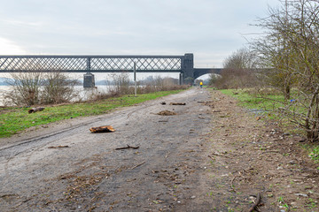 A dirt bike path on the banks of a river in western Germany after a flood, visible trash, tree branches and rail bridge.