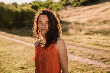 Fototapeta premium Beautiful young girl with dark curly hair in bright orange dress walks through an autumn forest or field at sunset. Woman is smiling and happy. Romantic mood.