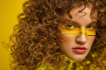 Close up fashion studio portrait of young beautiful women with curly hair. Nice girl with afro hair and bright make up wearing yellow eyeglasses over yellow background. Beauty and hair care concept.