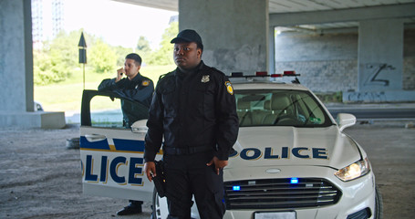 Serious handsome african american police officer looking around standing by patrol car. Two young policemen on duty keep guard on city at daytime. © Fractal Pictures