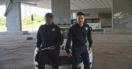 Confident brutal handsome men police officers getting out leaning on car looking on camera with serious expression. Police patrol the city. City security.