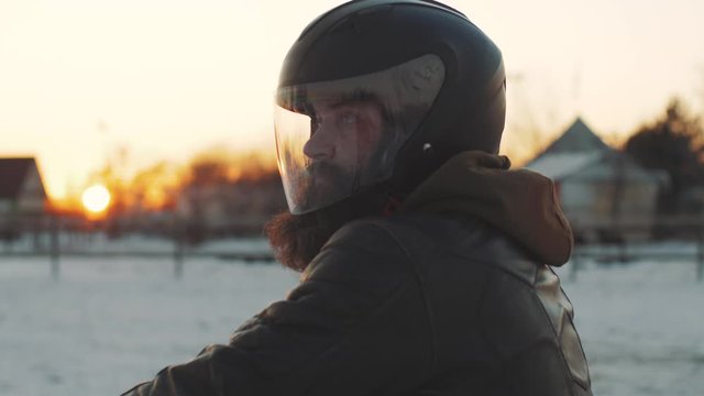 Portrait of a man motorcyclist in a black helmet at sunset on snow field. Man rides a motorcycle in cold winter