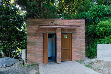 A small building in a park with two doors. The toilet in the botanical garden is laid out of brick on a background of trees. Red brick building in a park area with an open door.