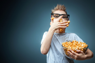 The boy holds in his hands and eats popcorn watching a movie in 3D glasses, blue background. The concept of a cinema, films, emotions, surprise, leisure. streaming platforms.