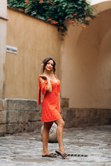 Beautiful girl in red dress posing on city streets. Pretty brunette posing against building background. Stunning young woman walking in the city