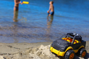 Fototapeta na wymiar Children toy car jeep of black and yellow color on sand near the water on the beach of blue sea. Boys play water guns. Vacation with kids on coast on warm sunny summer day. Tourism in Europe,Slovenia