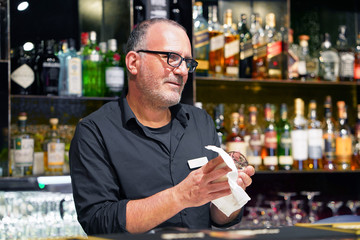 Elderly bartender with eyeglasses cleans the glass. A handsome bartender polishes a glass of wine glasses. The concept of service. 