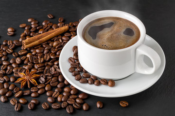 Hot coffee in white elegant cup with saucer on a background of roasted coffee beans over a black stone serving board. Coffee with cinnamon and anise.
