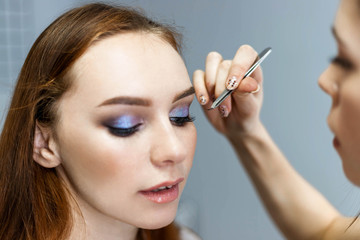 Obraz premium Skillful visagiste in white t-shirt is holding brush, applying make-up for young model while posing sideways against gray studio background. Close up