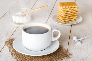Hot coffee in white elegant cup, stack of sweet square cookies, sugar and white smartphone headphones on a white wooden table. Steaming hot morning beverage and pastries.