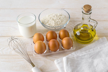 Raw ingredients prepared for cooking omelette: six brown eggs, glass of milk, some flour and salt, olive oil on a white wooden table. Easy preparation of breakfast.
