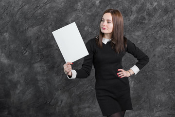 Brunette woman in black dress, hand on hip, holding white empty blank board ready for your text or product, posing on gray background. Close up