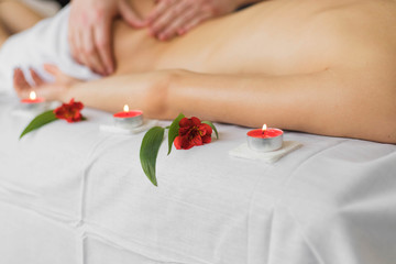 Hands of masseur making therapeutic back massage for female lying on couch in private clinic. Flowers and burning candles are nearby. Close up