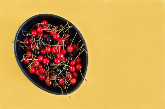 Black ceramic bowl with wild cherries on yellow background. Copy Space