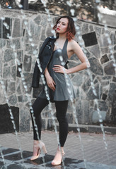 A woman dressed in a fashionable leather jacket, a silver dress and leggings, surrounded by spray of a fountain.