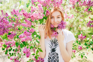 Obraz na płótnie Canvas Handsome woman with red hair and pretty smile posing in lilac garden with flowers and enjoys the smell