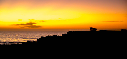 summer beach coast during sunset with a retro camper car standing on the cliff in the shadow of sun going down
