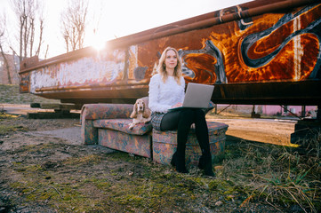 Cheerful caucasian young freelancer girl sitting on old couch outdoor over rusty graffiti wall on...