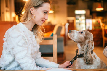 Young blonde girl in white sweater caring her lovely cocker spaniel puppy at the table in caffe.