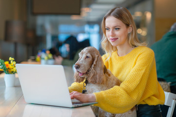 Young caucasian blonde freelancer girl holding her cocker spaniel dog on her knees in co working space and using laptop while working or studying.