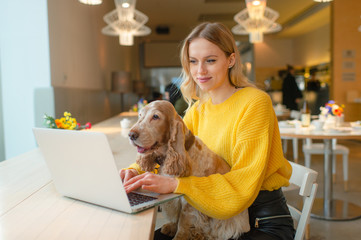 Young caucasian blonde freelancer girl holding her cocker spaniel dog on her knees in co working space and using laptop while working or studying.