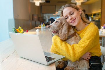 Happy smiling blonde woman hugging her lovely cocker spaniel puppy with eyes closed and joyful face expression in front of laptop on the table in caffe.