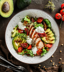 Healthy salad with fillet chicken, avocado, tomatoes, lettuce, greens, pine nuts and sauce in bowl on wooden background. Healthy food, clean eating, dieting, Buddha bowl, top view