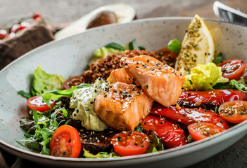 Healthy salad with fillet salmon, quinoa, avocado sauce, grilled pepper, tomatoes, lettuce, arugula in plate on wooden background. Healthy food, clean eating, Buddha bowl, dieting, close up
