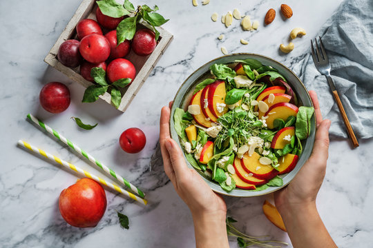 Hands holding fresh summer salad with peach, spinach, micro greens, plums, feta cheese and almonds on light marble background. Healthy food, clean eating, Buddha bowl salad, top view