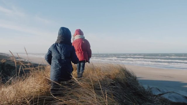 Little girl and boy child in warm coats walk along tall grass dunes at amazing sunny windy winter sea beach slow motion.