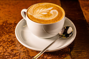 A Cup of cappuccino with a pattern on the foam is on the table in the early morning, while it is still dark.
