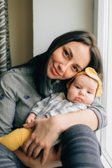 Portrait of beautiful woman with black hair sits on a windowsill with her pretty little child and smiles