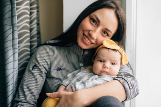 Portrait of beautiful woman with black hair sits on a windowsill with her pretty little child and smiles