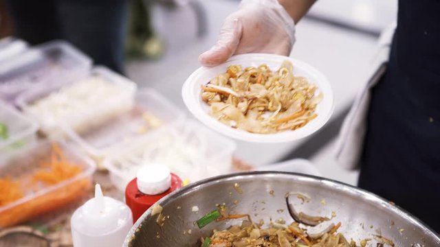 Close up of cook hands putting asian noodles into the disposable plate, asian street food concept. Art. Chef in cooking gloves preparing food outdoors.