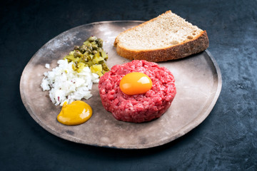 Gourmet tartar raw from beef fillet with yellow of the egg, gherkin and farmhouse bread as closeup on a modern design rustic plate