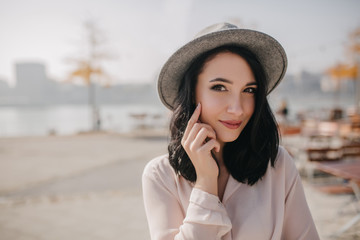 Good-looking brunette lady in hat playfully touching her face. Outdoor portrait of elegant young woman standing on blur nature background with sincere smile.