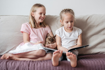 Two little girls sitting barefoot on the couch with Burmese cat watching a book