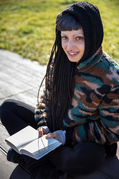 Portrait of smiling young woman with piercings and braids sitting on bench with a book, Como, Italy