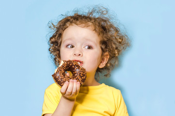 little curly girl eats, bites a delicious chocolate donut with an appetite and with pleasure, on blue background. trend. human emotions in food