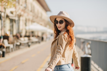 Portrait of laughing ginger woman looking over shoulder while walking down the street. Photo of joyful red-haired lady in sunglasses chilling at embankment.