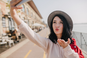 Obraz premium White female model sending air kiss while posing in sunny day. Outdoor portrait of spectacular pale girl making selfie during walk around city.