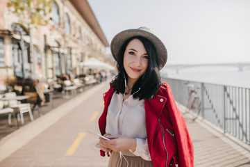 Obraz premium Adorable girl with straight short hair listening music at embankment in sunny day. Outdoor photo of elegant caucasian female model expressing positive emotions near street cafe.